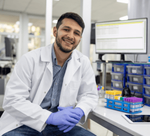 Scientist in a lab coat and gloves seated at a workstation with test tube and a tablet